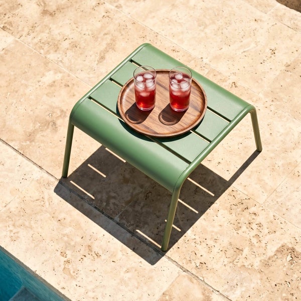 Grüner Gartenbeistelltisch aus Metall mit Lattenstruktur auf einer Steinterrasse am Pool mit Getränken auf einem Holztablett.