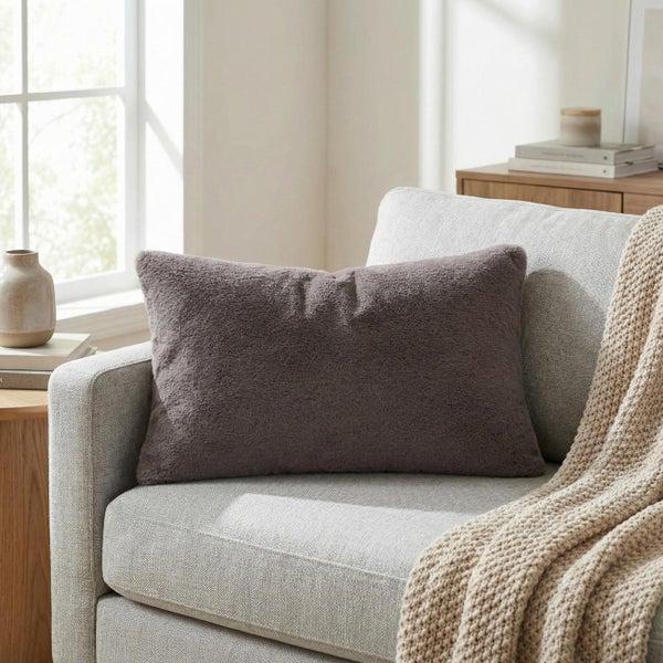 Rechteckiges Dekokissen mit weicher Kunstfell-Textur auf einem hellgrauen Sessel in einem hellen Wohnzimmer.
