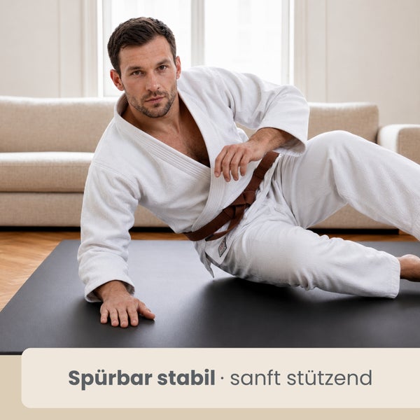 Ein Mann in einem weißen Kampfanzug stützt sich auf einer stabilen, dunklen Bodenmatte in einem Wohnzimmer ab.