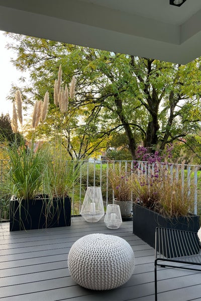 Moderne Terrasse mit grauen Dielen, schwarzen Pflanzkästen mit hohem Ziergras, weißem Strick-Pouf und dekorativen Windlichtern vor Bäumen.