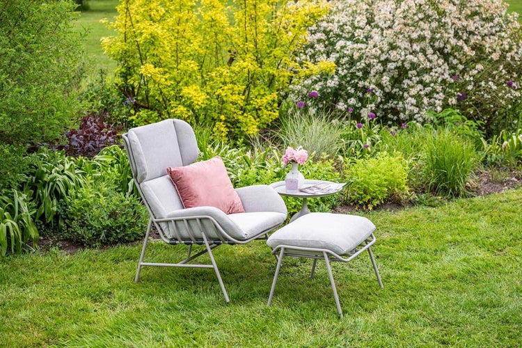 Moderner Loungesessel mit Hocker und Beistelltisch in einer Gartenanlage mit Blumen und Sträuchern.