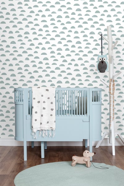 Ein Kinderzimmer mit einem hellblauen Kinderbett, einer Spielzeugfigur und einer Dekoration an einem Kleiderständer vor einer gemusterten Tapete.