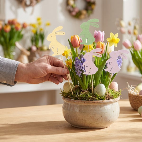 Hand dekoriert einen Blumentopf mit Frühlingsblumen, Moos, Ostereiern und hölzernen Osterhasensteckern als festliche Osterdekoration auf einem Tisch.
