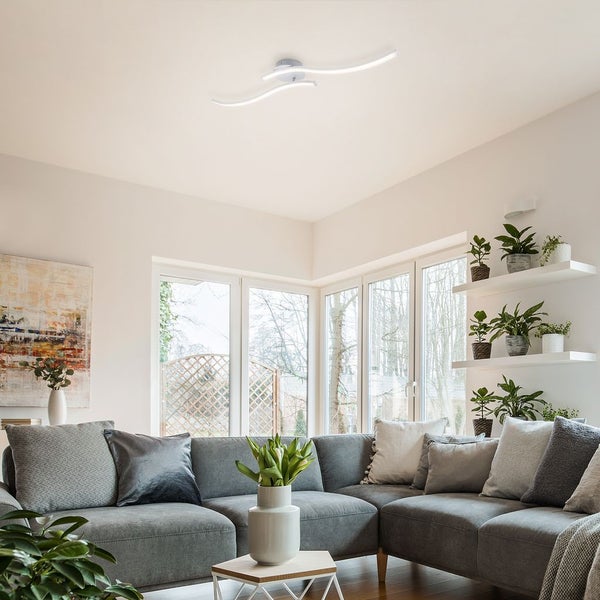 Wellenförmige LED-Deckenleuchte in einem hellen, modern eingerichteten Wohnzimmer mit großem grauem Ecksofa und Zimmerpflanzen auf Regalen.
