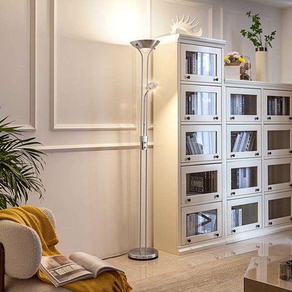 Moderner Deckenfluter aus Metall mit Lesearm in einem hellen Wohnzimmer neben einem weißen Glasregal und einem Sessel mit Decke.