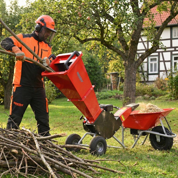 Ein Mann in Schutzkleidung füttert Äste in einen roten Hecht Gartenhäcksler, wobei die Holzschnitzel in eine Schubkarre fallen.