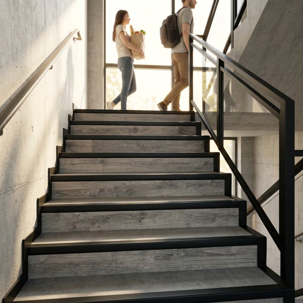 Moderne Treppe mit Bodenbelag in grauer Holzoptik, schwarzen Treppenprofilen aus Metall und Glasgeländer in industriellem Ambiente mit Betonwänden.
