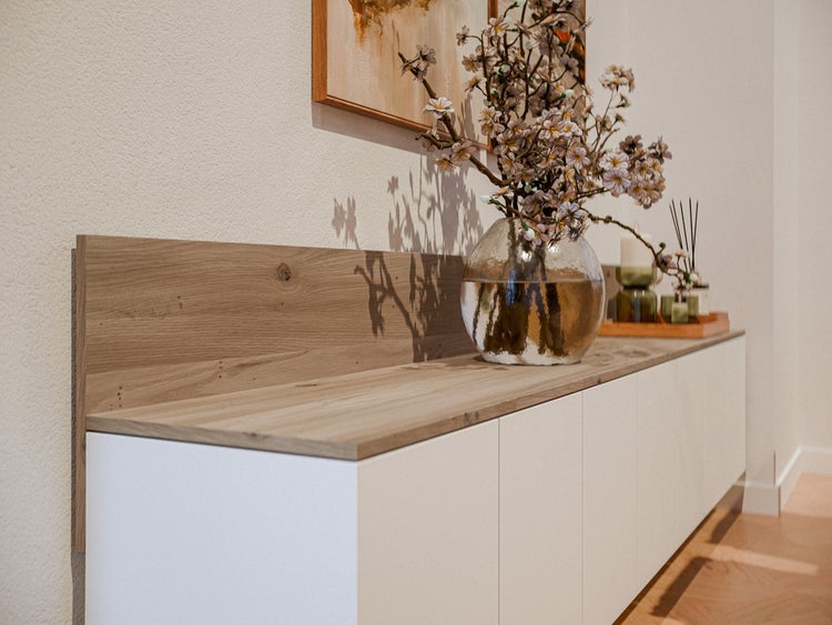 Sideboard mit Arbeitsplatte und Wandabschlussleiste in Eichenoptik sowie weißen Fronten in einem modern eingerichteten Raum.