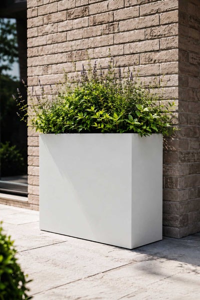 Moderner rechteckiger Pflanzkasten in Weiß vor einer Klinkerwand, bepflanzt mit grünen Pflanzen und violetten Blüten auf einer Terrasse.