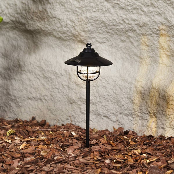 Klassische Außenwegeleuchte aus Metall mit Schirm und Glaskäfig in einem Beet mit Rindenmulch vor einer Steinmauer.