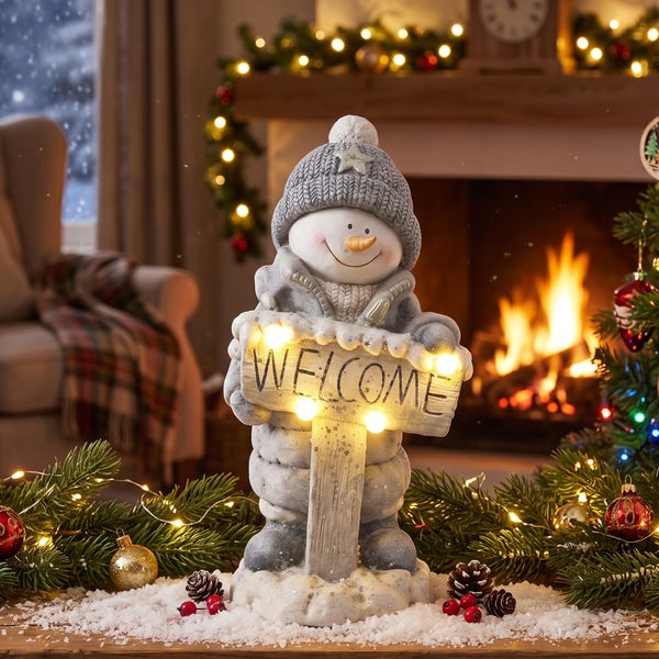 Weihnachtsdekoration Schneemann mit beleuchtetem Welcome Schild aus Steinzeug vor einem Kamin in einer gemütlichen winterlichen Wohnstube.