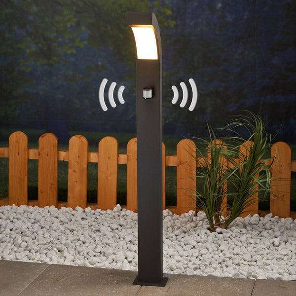 Moderne LED-Außenstehleuchte mit integriertem Bewegungsmelder, die einen Gartenbereich mit Kieselsteinen und einem Holzzaun beleuchtet.