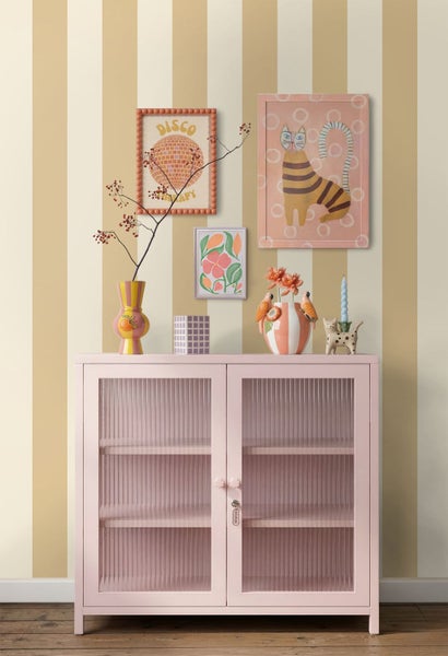 Ein rosa Metallschrank mit Riffelglastüren vor einer gestreiften Tapete. Dekorative Vasen und Wandbilder ergänzen den hellen Wohnbereich.
