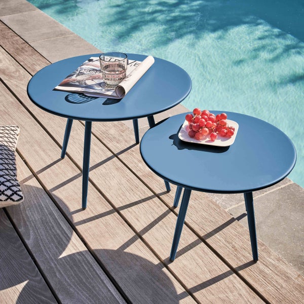 Zwei runde blaue Beistelltische aus Metall in verschiedenen Höhen auf einer Holzterrasse am Pool mit Magazin, Wasserglas und Weintrauben.