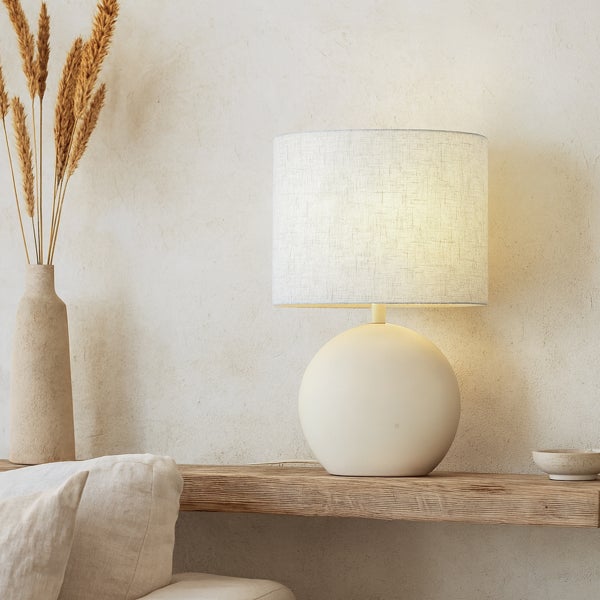 Tischleuchte mit rundem Keramikfuß und Stoffschirm in Zylinderform auf einem Holzregal neben einer Vase mit Trockengras.