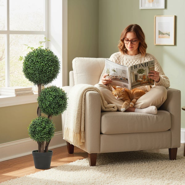 Künstlicher Buchsbaum mit drei Kugeln im schwarzen Topf in einem hellen Wohnzimmer neben einem Sessel mit einer lesenden Person.