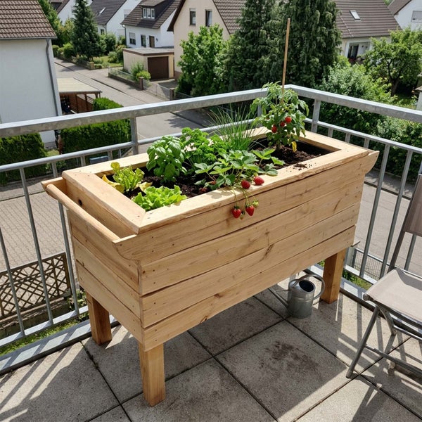 Ein hölzernes Hochbeet auf einem Balkon, bepflanzt mit frischem Gemüse und Kräutern wie Salat, Basilikum, Erdbeeren und Tomaten.