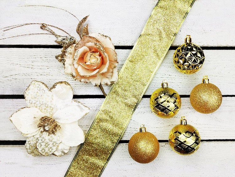 Weihnachtsdekoration bestehend aus zwei Kunstblumen, einem breiten Geschenkband und fünf glitzernden Christbaumkugeln auf weißem Holzuntergrund.