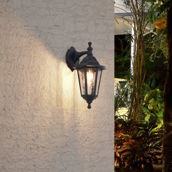Schwarze Außenwandleuchte in Laternenform mit strukturiertem Glas, montiert an einer hellen Außenwand im Gartenbereich.