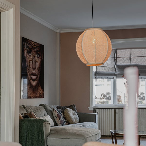 Eine runde Pendelleuchte aus geflochtenem Material in einem gemütlichen Wohnzimmer mit Sofa und Wandbild.