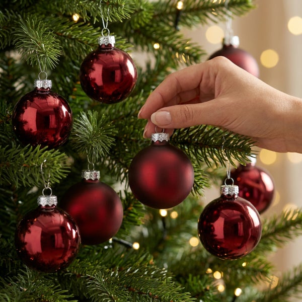 Eine Hand platziert eine matte Weihnachtsbaumkugel aus Glas an einem Nadelbaum mit weiteren glänzenden Christbaumkugeln.