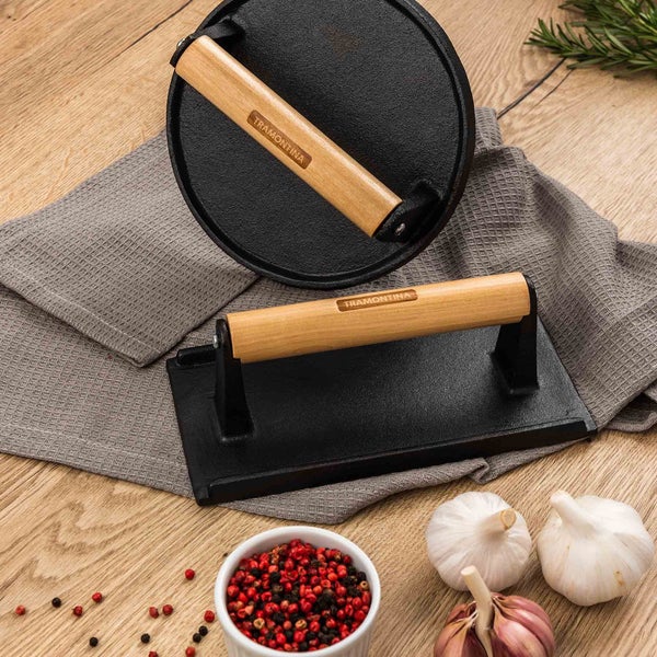 Zwei Grillpressen aus Gusseisen mit Holzgriffen der Marke Tramontina auf einem Küchentuch, zusammen mit einer Schale gemischter Pfefferkörner und Knoblauchzehen.