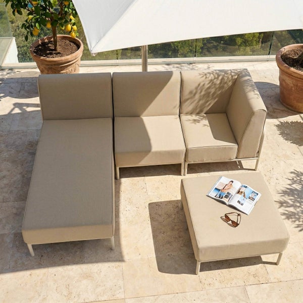 Modulares Garten-Lounge-Set aus gepolsterten Elementen und einem Hocker auf einer hellen Steinterrasse mit Sonnenschirm und Pflanzen.