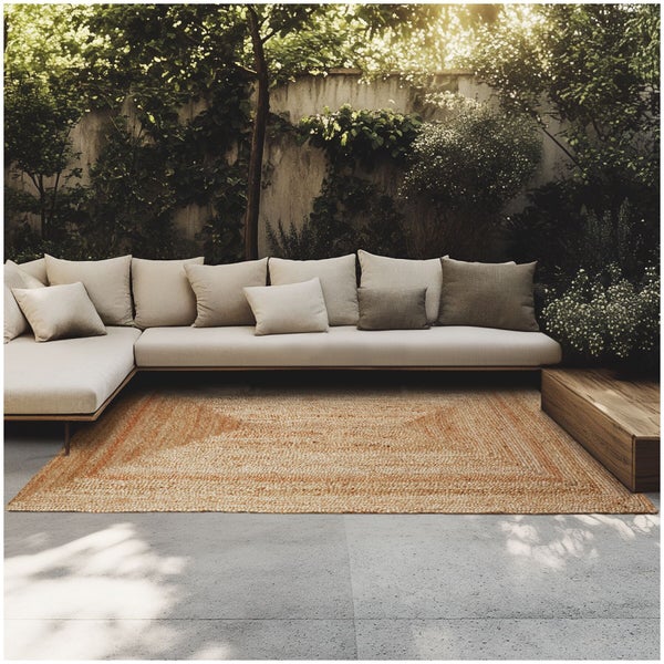 Rechteckiger Jute Teppich vor einem Ecksofa auf einer Terrasse in einem Garten mit Pflanzen und einer Mauer.