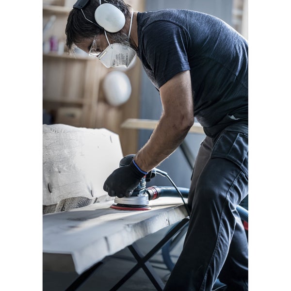 Ein Mann schleift Holz mit einem Exzenterschleifer und trägt dabei Schutzausrüstung wie Gehörschutz, Schutzbrille und Atemschutzmaske.