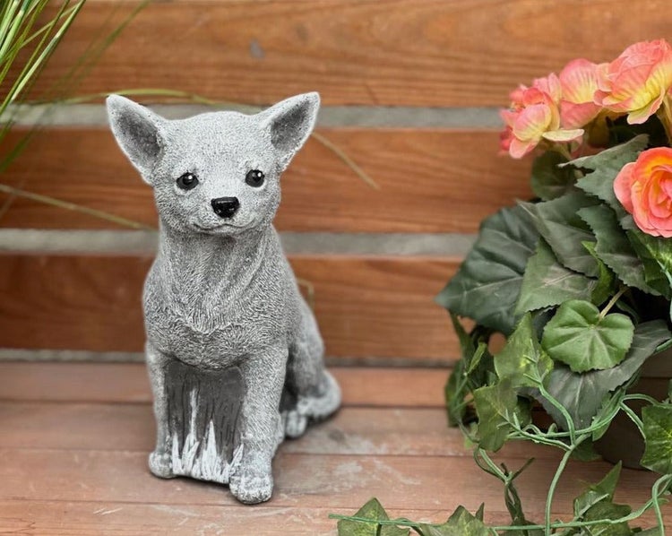 Gartenfigur eines sitzenden Chihuahuas aus Steinguss neben einer Pflanze vor einer Holzwand.