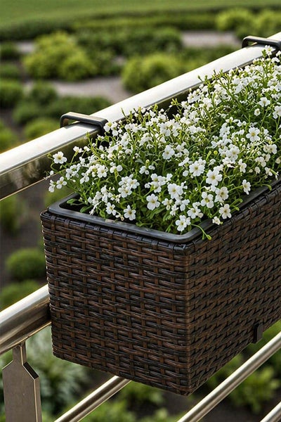 Balkonkasten in Rattan-Optik mit weißen Blumen an einem Metallgeländer befestigt.