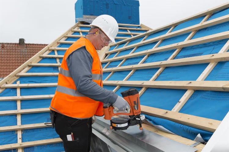 Handwerker mit Schutzhelm und Warnweste arbeitet auf einem Dach mit einer Druckluftnagelpistole bei der Befestigung einer Metallrinne.