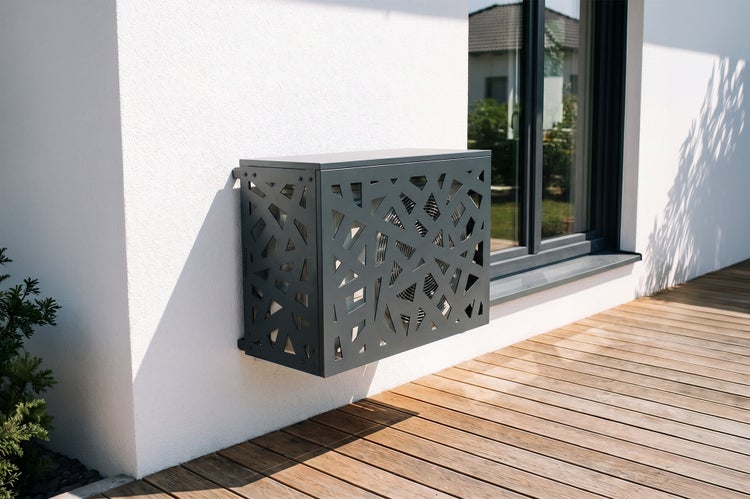 Wandmontierte Klimaanlagen-Verkleidung aus Metall mit geometrischem Muster an einer weißen Hauswand neben einem Fenster über einer Holzterrasse.