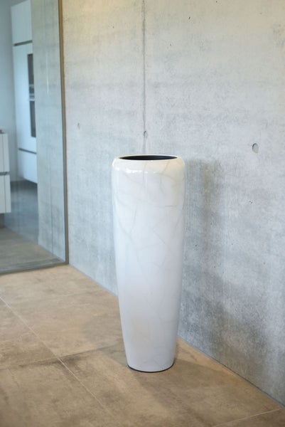 Hohe, glänzende Bodenvase in Weiß mit feiner Marmorierung vor einer Betonwand in einem modernen, hellen Innenraum.
