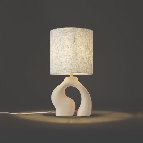 Beige Tischleuchte mit skulpturalem Fuß und hellem, zylindrischem Textilschirm in Leinenoptik.