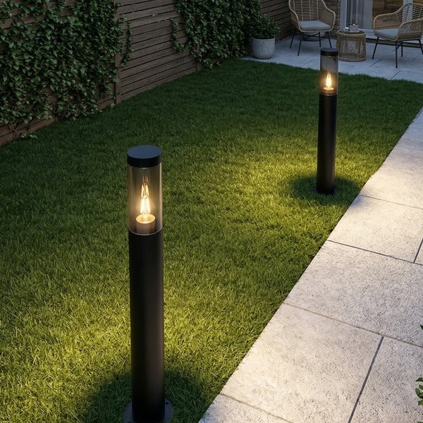 Zwei schwarze zylinderförmige Außenstehleuchten mit warmen Filament-Leuchtmitteln beleuchten einen Rasen neben einem gepflasterten Gartenweg.