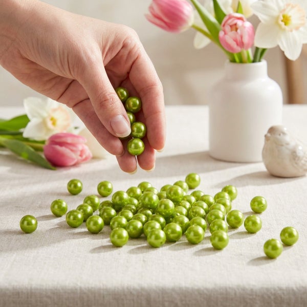 Hand haelt kleine gruene Schmuckperlen fuer Dekorationen und Bastelarbeiten auf einem Tisch mit Blumen im Hintergrund