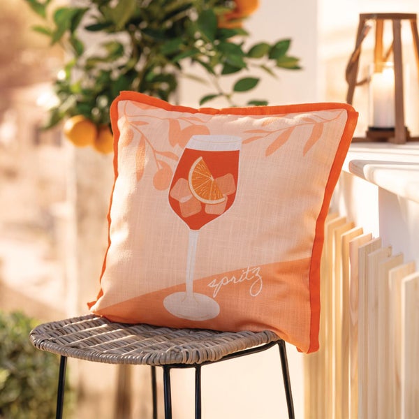 Dekokissen mit Spritz-Motiv, Glas mit Orangenscheibe und Eiswürfeln, auf einem geflochtenen Gartenstuhl im Außenbereich.