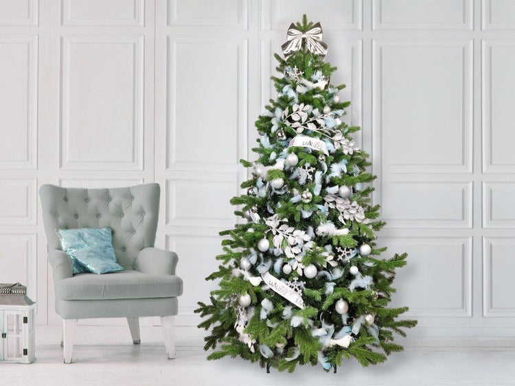 Geschmückter Weihnachtsbaum mit silbernen und hellblauen Dekorationen neben einem Sessel in einem hellen, festlich eingerichteten Raum.