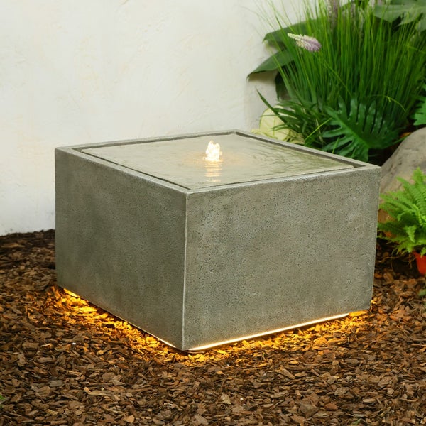 Quadratischer Gartenbrunnen in Betonoptik mit sprudelndem Wasser und LED-Bodenbeleuchtung auf Rindenmulch.