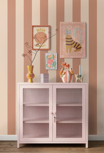 Moderner Innenraum mit einem rosa Metallschrank mit Riffelglastüren vor einer gestreiften Tapete. Auf dem Schrank stehen dekorative Vasen.