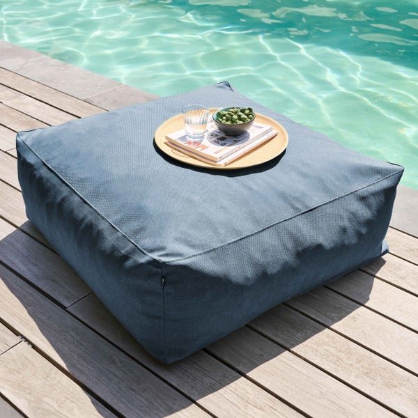 Großer quadratischer blauer Outdoor-Pouf auf einer Holzterrasse am Pool, genutzt als Beistelltisch mit einem Tablett.