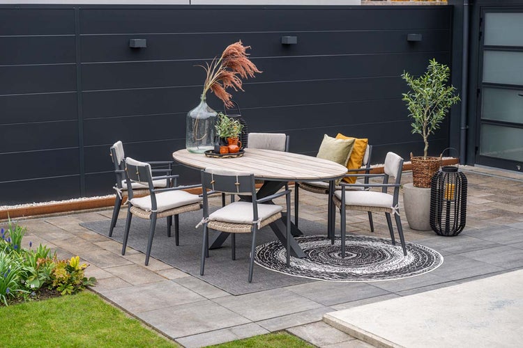 Moderne Gartenloungegruppe mit ovalem Holztisch und gepolsterten Gartenstühlen auf einer Terrasse vor einer dunklen Sichtschutzwand.