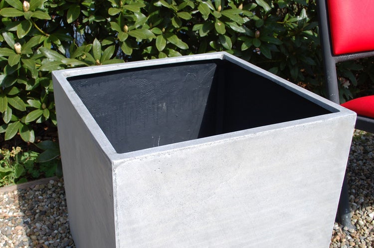 Quadratischer Pflanzkübel in Betonoptik im Garten auf Kies vor Sträuchern.