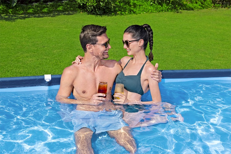 Ein Paar sitzt mit Getränken in einem blauen Gartenpool in einem hellen Garten mit Rasenfläche.