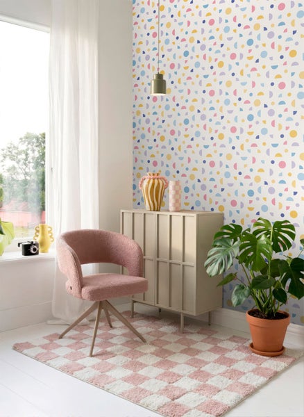 Helles Zimmer mit bunter Tapete mit geometrischen Formen, einem rosa Sessel, beigem Sideboard und einer Monstera Pflanze auf kariertem Teppich.