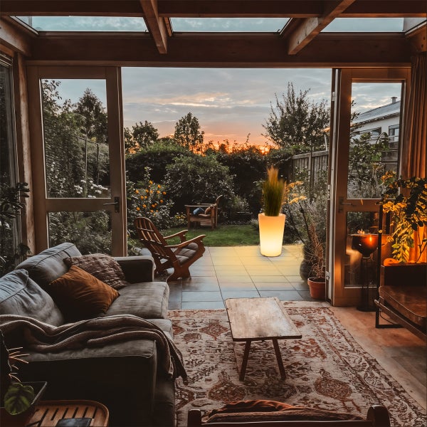 Gemütlicher Wohnraum mit Sofa und Teppich, Blick durch Glastüren auf Terrasse mit beleuchtetem Pflanzkübel und Garten bei Sonnenuntergang.