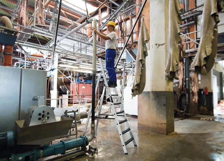 Ein Arbeiter steht auf einer stabilen Aluminium Stehleiter in einer industriellen Fertigungshalle und führt Wartungsarbeiten an Rohrleitungen aus.