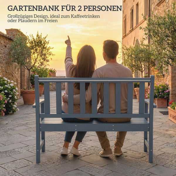 Eine Gartenbank für zwei Personen auf einer Terrasse bei Sonnenuntergang, auf der ein Paar sitzt und in die Ferne deutet.