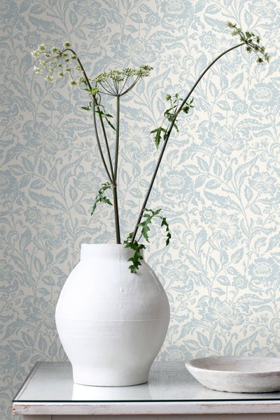 Vliestapete mit blauem floralem Muster in einem hellen Raum mit weißer Keramikvase und Pflanze.